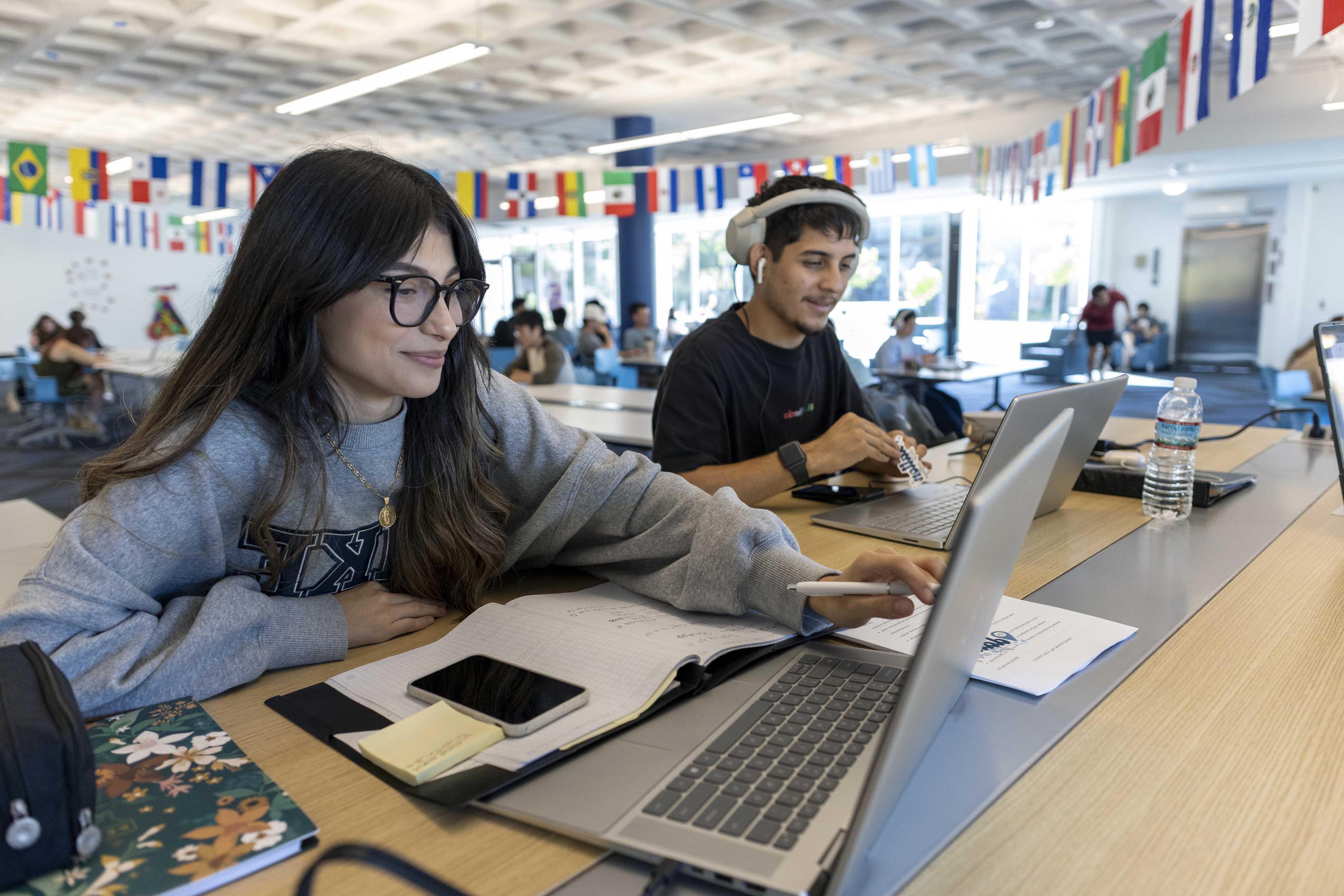 Students working on laptops in a room with international flags along the ceiling